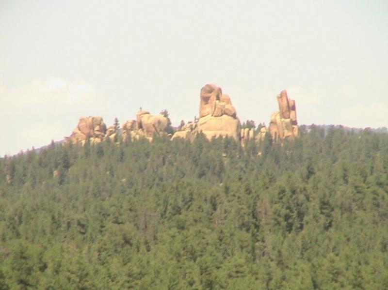 A rocky outcrop rises above a dense forest, featuring unique rock formations against a clear sky. The landscape showcases natural beauty and rugged terrain. Chair Rocks mountain bike trail.