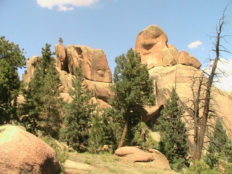A scenic view of large rock formations surrounded by greenery, featuring tall trees and a blue sky with scattered clouds. The rocky landscape showcases unique shapes and textures, creating a natural outdoor setting. Chair Rocks mountain bike trail.