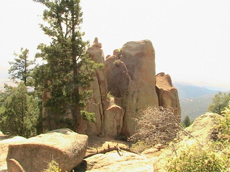 Rock formations surrounded by coniferous trees, with a mountainous landscape visible in the background under a bright, hazy sky. Chair Rocks mountain bike trail.