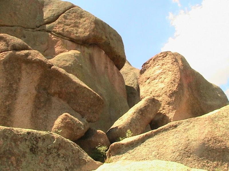 A close-up view of large, weathered rock formations under a bright blue sky. The rocks have various shapes and textures, showcasing their natural ruggedness, with some areas displaying green vegetation peeking through the crevices. Chair Rocks mountain bike trail.