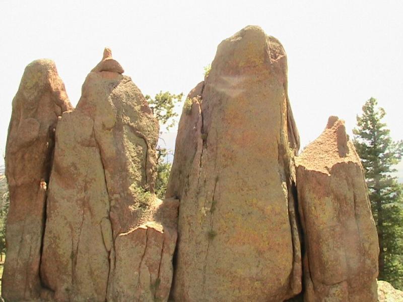 A group of large, natural rock formations with rugged surfaces and varying heights, surrounded by trees in the background. The scene is well-lit, showcasing the textures and colors of the rocks. Chair Rocks mountain bike trail.