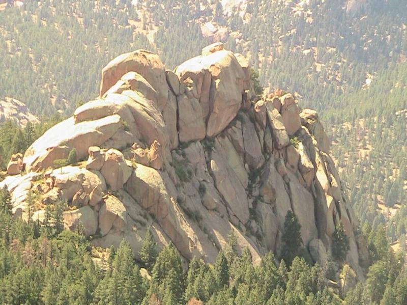 A rocky mountain formation with multiple large boulders, surrounded by dense green forest and a backdrop of distant hills. The scene captures natural textures and colors under clear skies. Chair Rocks mountain bike trail.