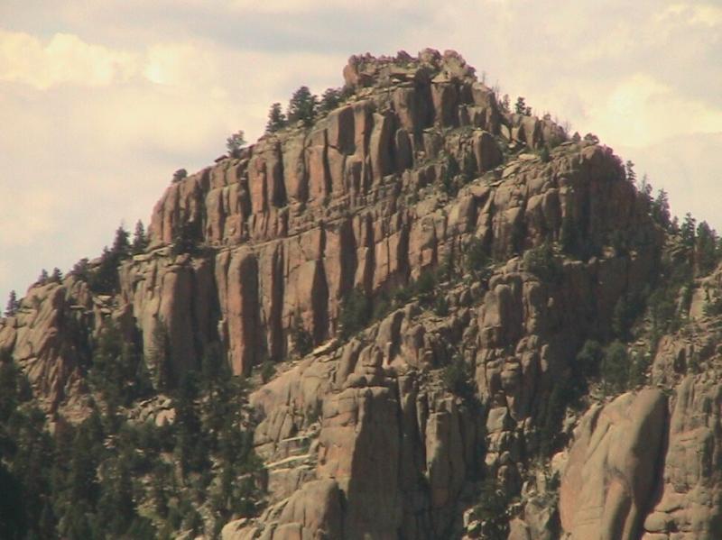 A rugged mountain peak with steep, jagged rock formations and scattered pine trees, set against a cloudy sky. Chair Rocks mountain bike trail.