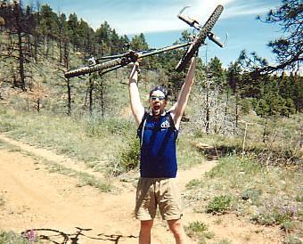 A person wearing a tank top and shorts is standing on a dirt trail in a forested area, triumphantly holding a mountain bike above their head. They are smiling and raising their arms in celebration. Chair Rocks mountain bike trail.