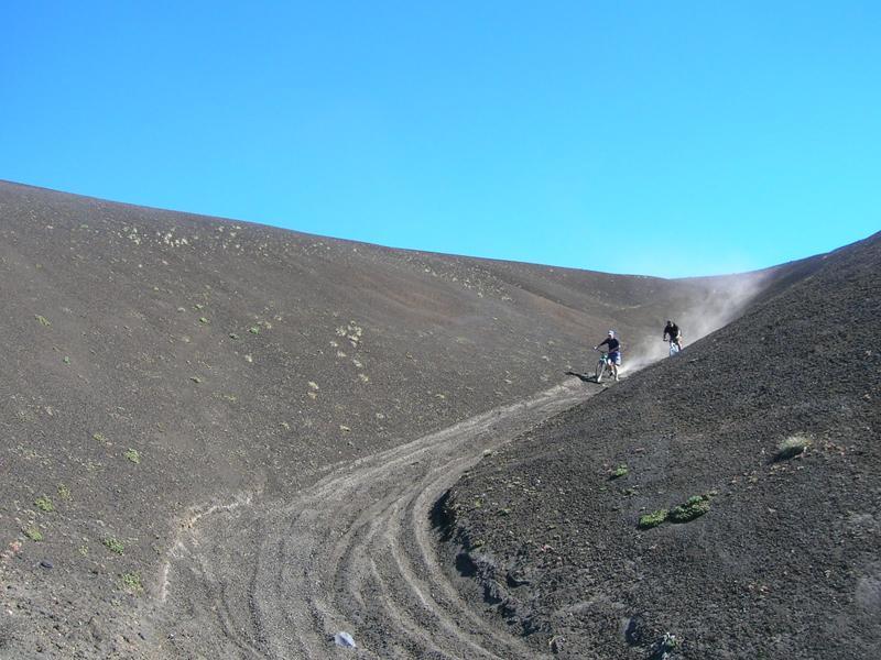 Two mountain bikers navigate a winding dirt trail on a steep volcanic hillside, with a clear blue sky above and dust trailing behind them. The landscape features a mix of dark volcanic soil and patches of green vegetation. Parque Huilo Huilo- Choshuenco Volcano mountain bike trail.