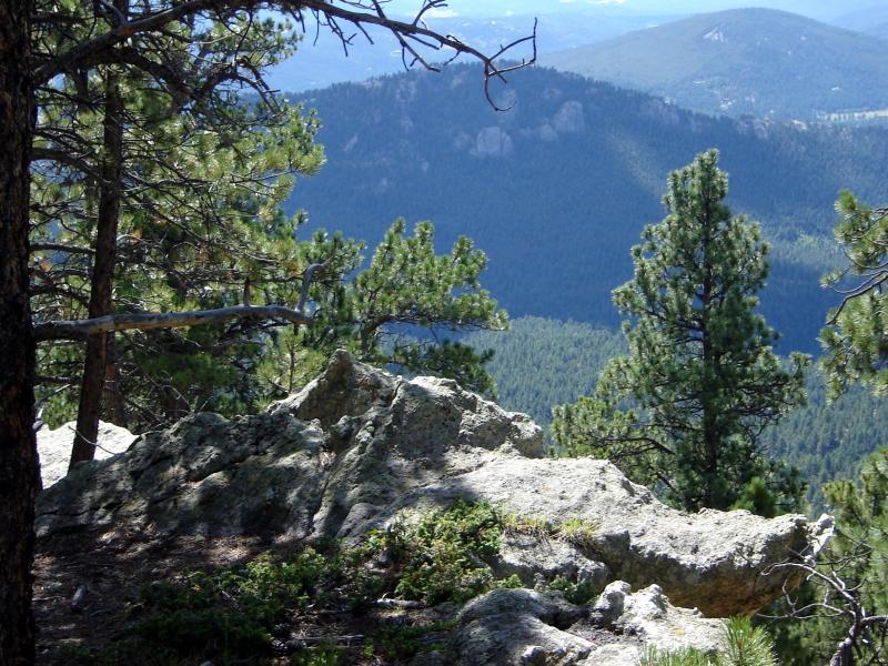 A rocky outcrop in the foreground with pine trees surrounding it, overlooking a vast mountain landscape dotted with greenery under a clear blue sky. Bergen Peak mountain bike trail.