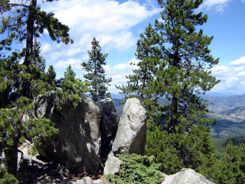 Rocky outcrop surrounded by evergreen trees with a scenic view of mountain ranges and a blue sky filled with clouds. Bergen Peak mountain bike trail.