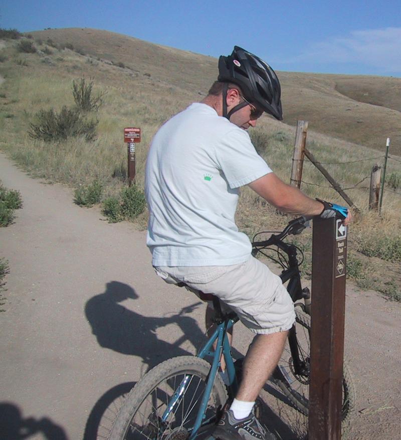A cyclist in a helmet pauses on a mountain bike near a trail marker on a dirt path, surrounded by grassy hills under a clear blue sky. The cyclist is wearing a light blue shirt and shorts, and is looking at the trail sign, which indicates the direction of the bike trail. Shane's Loop mountain bike trail.