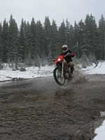 A motorcyclist performing a jump over a muddy water puddle in a snowy forest setting, surrounded by tall pine trees.