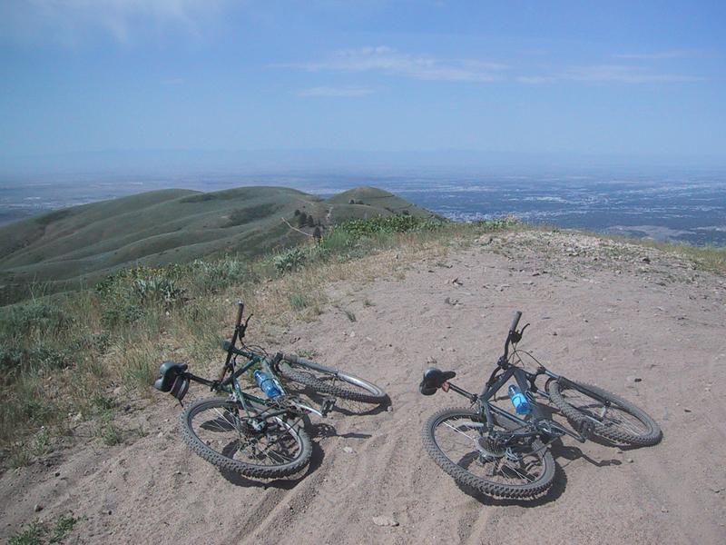 Two mountain bikes lying on sandy ground at the top of a hill, with a panoramic view of rolling green hills and a distant landscape under a clear blue sky. 8th Street Motorcycle Trail mountain bike trail.