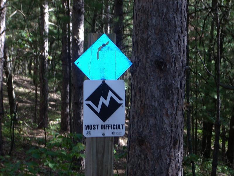 A trail sign in a wooded area indicating a difficult path. The sign features a blue diamond shape at the top, followed by a black diamond symbol and the text "MOST DIFFICULT" on a white background. Surrounding trees provide a natural backdrop. Standing Rocks mountain bike trail.