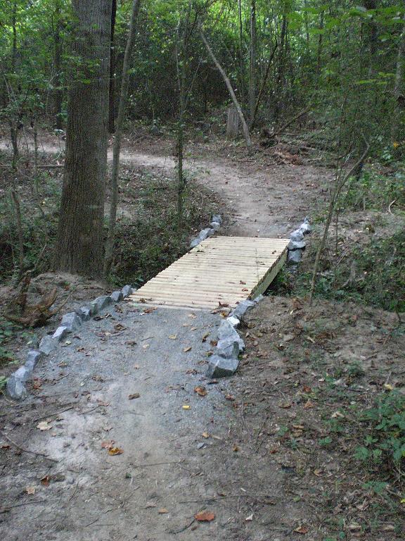 A wooden bridge supported by stone edges spans a small creek in a forested area. Surrounding the bridge are trees and underbrush, with dirt paths leading away from it, suggesting a hiking trail. Fallen leaves and natural debris are scattered along the ground, indicating a serene, natural environment. Jetton Park mountain bike trail.