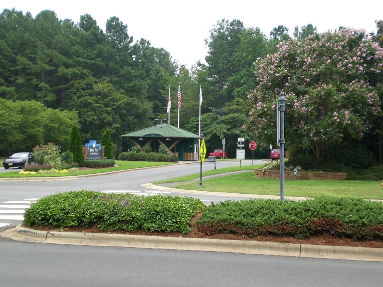 A landscaped entrance with a green canopy over the guard house, surrounded by trees and flowering shrubs. Flags are flying in front of the building, and vehicles are parked nearby. The setting is peaceful, showcasing well-maintained greenery and a welcoming atmosphere. Jetton Park mountain bike trail.