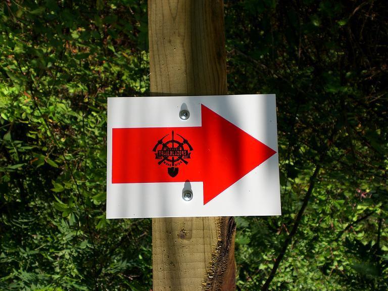 A white sign with a large red arrow pointing to the right, featuring a black logo that reads "Trailblazers" with an emblem design, mounted on a wooden post surrounded by greenery. Jetton Park mountain bike trail.