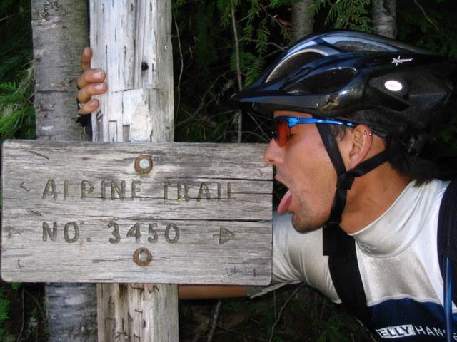 A person wearing a helmet and sunglasses playfully poses with a wooden trail sign that reads "ALPINE TRAIL NO. 3450," leaning towards it and sticking out their tongue. The background features trees, suggesting an outdoor setting. Alpine Trail mountain bike trail.