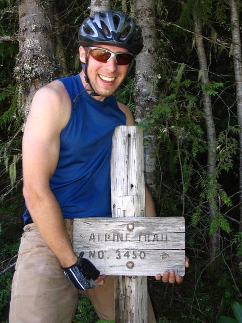 A smiling person wearing a bike helmet and sunglasses stands next to a wooden signpost labeled "Alpine Trail No. 3450," surrounded by trees. The person is dressed in a sleeveless blue top and is positioned playfully next to the sign. Alpine Trail mountain bike trail.