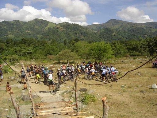 A large group of cyclists gathered in a grassy area surrounded by lush green mountains under a partly cloudy sky. Some cyclists are on bicycles, while others are standing near them. There is a rustic wooden bridge in the foreground leading to the group. Cordillera Central mountain bike trail.