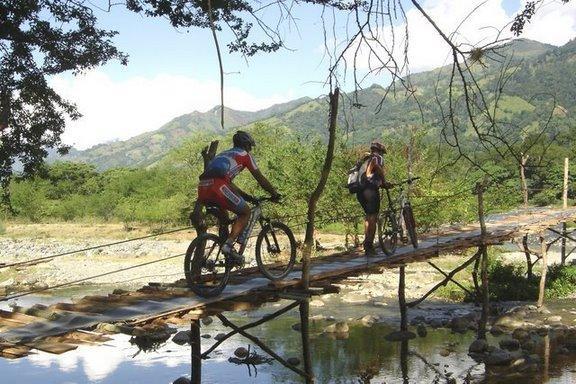 Two mountain bikers crossing a rustic wooden bridge over a shallow river, surrounded by lush greenery and mountains in the background. The scene captures the adventurous spirit of outdoor cycling in a natural setting. Cordillera Central mountain bike trail.