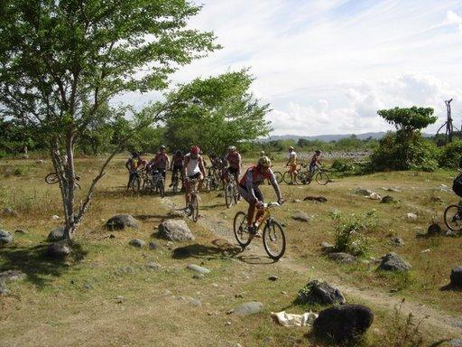 A group of mountain bikers navigating a rocky, outdoor trail surrounded by trees and greenery on a sunny day. Cordillera Central mountain bike trail.