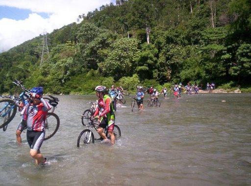A group of cyclists wade through a shallow river while carrying their bicycles, surrounded by lush greenery and trees. The scene captures the adventurous spirit of outdoor biking, with clear skies above and a varied landscape in the background. Cordillera Central mountain bike trail.