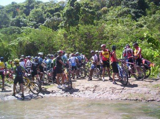 A large group of cyclists gathered by a riverbank, surrounded by lush greenery. The cyclists, wearing helmets and colorful attire, are gathered around their bikes, some standing and others sitting. The atmosphere appears lively and social, with the sunlight filtering through the trees. Cordillera Central mountain bike trail.