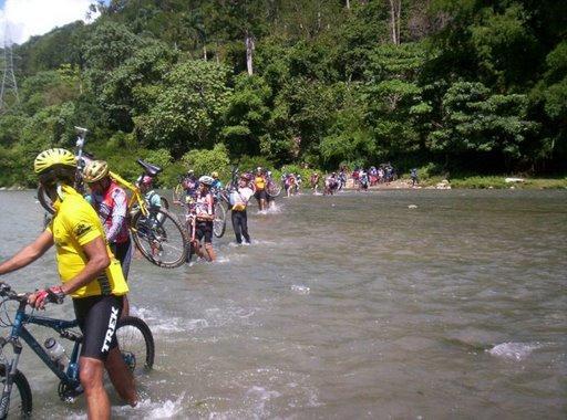 A group of cyclists crossing a shallow river while carrying their bikes, surrounded by lush greenery. Some cyclists are wading through the water, and others are walking along the shoreline. The scene illustrates a recreational outdoor event. Cordillera Central mountain bike trail.