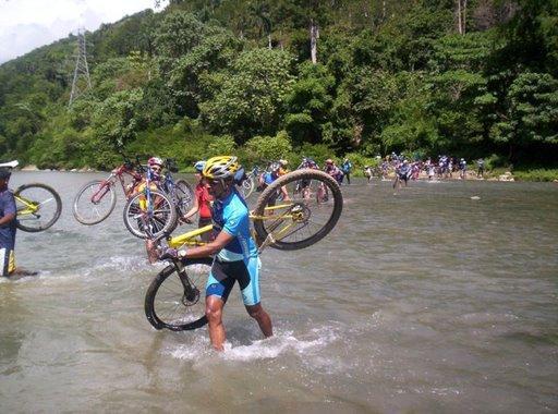A group of cyclists wading through a shallow river while carrying their bikes. Lush greenery and trees are visible in the background, with some cyclists walking alongside the water. The weather appears sunny and bright. Cordillera Central mountain bike trail.