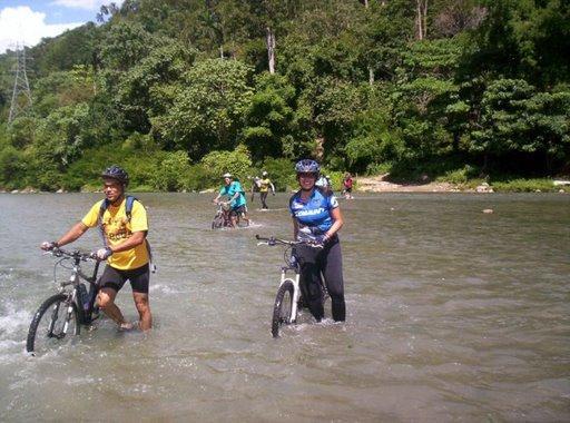 A group of cyclists wading through shallow water with their bikes. The scene is set in a lush, green landscape, with trees and hills in the background. Two cyclists in the foreground are walking alongside their bicycles, while others are visible further back in the water. Cordillera Central mountain bike trail.