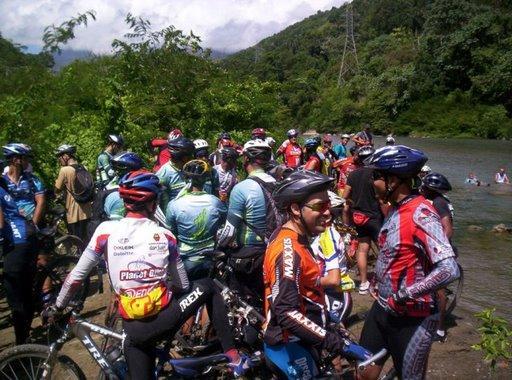 A group of mountain bikers gathered by a river, wearing cycling helmets and colorful jerseys. They are chatting and relaxing in a lush green outdoor setting, with trees and hills in the background. Some participants are near the water, while others are standing in clusters on the shore. Cordillera Central mountain bike trail.