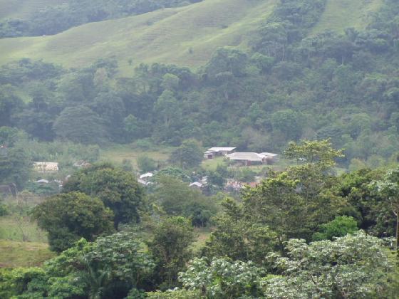 A lush green landscape with rolling hills in the background and a small cluster of buildings nestled among trees in the foreground. The scene is filled with various shades of green from the vegetation, creating a serene, natural setting. Cordillera Central mountain bike trail.