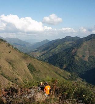 Two people are seen sitting on a grassy hilltop, overlooking a vast landscape of rolling green mountains under a clear blue sky with scattered clouds. Cordillera Central mountain bike trail.