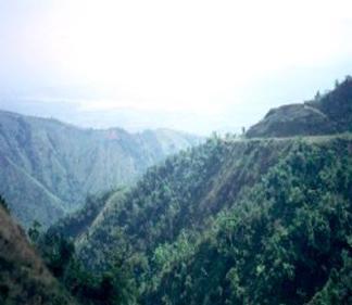A scenic landscape featuring rolling hills covered in lush greenery, with a misty sky in the background. The hills create a sense of depth and tranquility, highlighting the natural beauty of the area. Cordillera Central mountain bike trail.