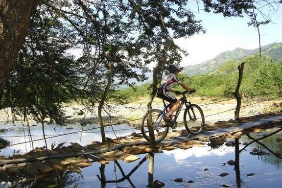 A cyclist riding a mountain bike across a narrow wooden bridge over a calm river, surrounded by lush greenery and mountains in the background on a sunny day. Cordillera Central mountain bike trail.