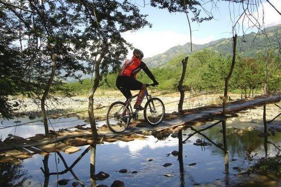 A cyclist riding a mountain bike across a wooden bridge over a stream, surrounded by lush greenery and mountains in the background. The scene captures an adventurous outdoor setting with clear blue skies and reflections in the water. Cordillera Central mountain bike trail.