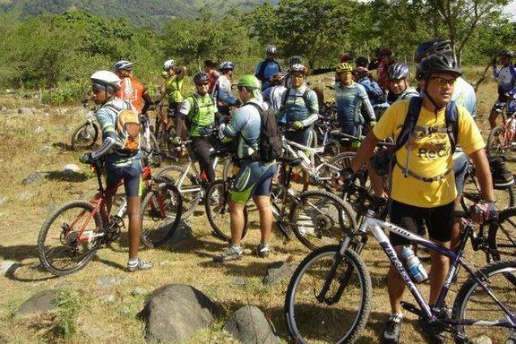 A group of mountain bikers gathered in a wooded area, some on their bikes and others standing nearby. They are wearing cycling gear and helmets, with various bicycles parked on the ground. The background features greenery and rocky terrain, suggesting a scenic outdoor location suitable for biking. Cordillera Central mountain bike trail.