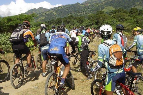 A group of mountain bikers is gathered on a dirt path surrounded by greenery and mountains. The cyclists, dressed in various colorful jerseys and helmets, are lined up and appear to be preparing for a ride or event. The landscape features lush hills in the background under a partly cloudy sky. Cordillera Central mountain bike trail.