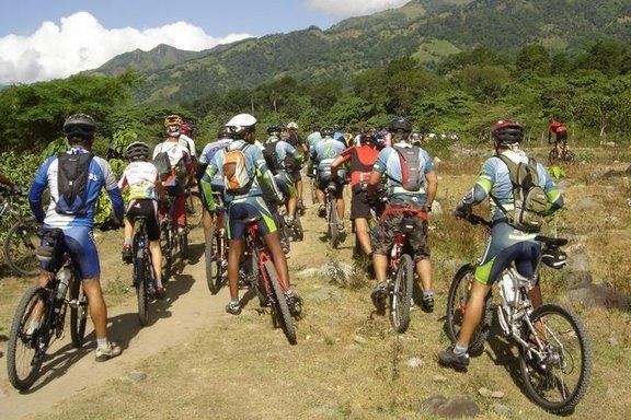 A group of mountain bikers riding on a dirt path through a lush landscape, with green mountains in the background and a clear sky. The cyclists are dressed in colorful jerseys and biking gear, heading into the scenic area. Cordillera Central mountain bike trail.