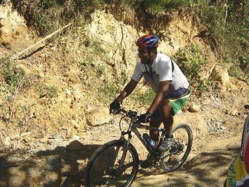 A person riding a mountain bike along a dirt path in a forested area, wearing a helmet and casual athletic clothing. Cordillera Central mountain bike trail.