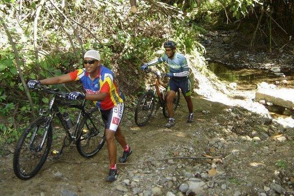 Two cyclists navigate a rugged trail, pushing their mountain bikes over a rocky path. The first cyclist, wearing a colorful jersey and sunglasses, walks with determination. The second cyclist, dressed in a blue shirt and black shorts, follows closely behind. Lush greenery surrounds the trail, with a stream visible in the background. Cordillera Central mountain bike trail.
