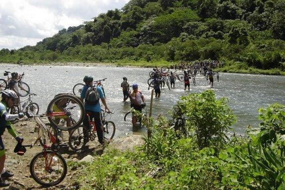 A group of cyclists is navigating a river crossing, some carrying their bikes while others wade through the water. Lush green vegetation surrounds the riverbank, and the sky is partly cloudy. Cordillera Central mountain bike trail.
