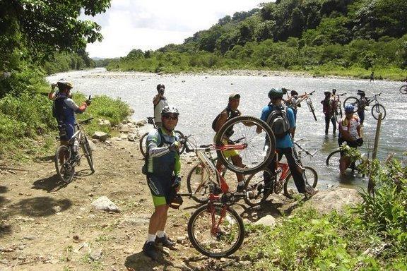 A group of mountain bikers standing by a riverbank, with some holding their bicycles and others wading in the shallow water. Lush green trees and a rocky shoreline are visible in the background, under a partly cloudy sky. Cordillera Central mountain bike trail.