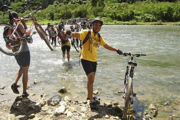 A group of mountain bikers fording a river, with some participants carrying their bicycles above their heads. One rider in a yellow shirt smiles while holding his bike, as others navigate the water in the background. Lush greenery surrounds the scene, indicating a natural setting. Cordillera Central mountain bike trail.
