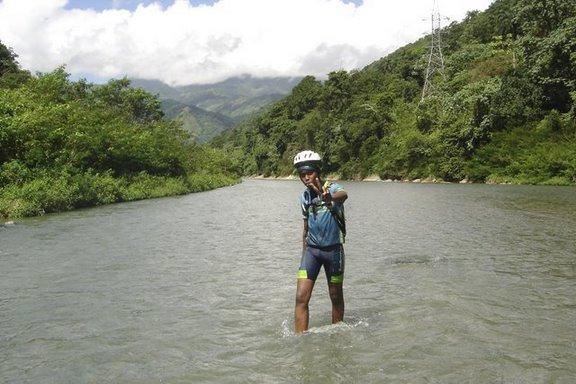 A person standing in a river with a thumbs-up gesture, wearing a bicycle helmet and sports clothing, surrounded by lush green vegetation and mountains in the background under a partly cloudy sky. Cordillera Central mountain bike trail.