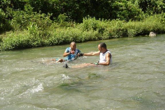 Two men are wading in a river, with water up to their waists. One man is reaching out to the other, who is kneeling in the water. The scene is surrounded by lush green vegetation, suggesting a natural, outdoor setting. The sunlight is bright, indicating a warm day. Cordillera Central mountain bike trail.
