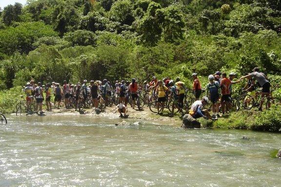 A large group of cyclists gathered along the edge of a river, with their bicycles lined up nearby. The scene is surrounded by lush green vegetation and trees, suggesting a tropical or subtropical environment. Some individuals appear to be interacting with the water, while others are standing and chatting. The atmosphere conveys a sense of community and outdoor adventure. Cordillera Central mountain bike trail.