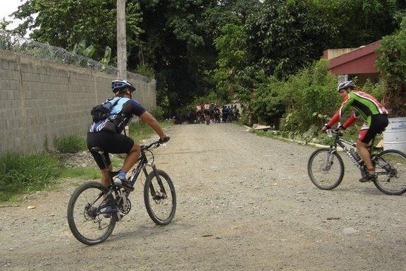 Two cyclists riding on a gravel road in a green, wooded area. One cyclist is wearing a blue and black outfit with a backpack, while the other is in a green and red outfit. Trees and a concrete wall line the path, with a group of people visible in the background. Cordillera Central mountain bike trail.