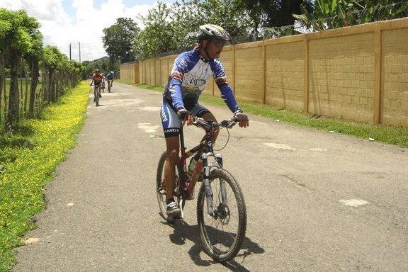 A cyclist in a blue and white jersey rides along a paved path lined with greenery and yellow flowers. Other cyclists can be seen in the background, enjoying a scenic ride on a sunny day. A brick wall runs alongside the path, and the sky is partly cloudy. Cordillera Central mountain bike trail.