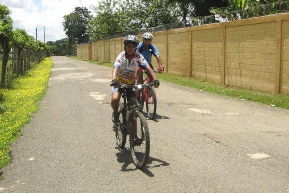 Two cyclists riding bicycles on a quiet, paved path lined with trees and a yellow wall. The sun is shining, and there are patches of green grass and yellow flowers along the edge of the road. One cyclist is dressed in a colorful jersey and wearing a helmet, while the other cyclist follows behind, wearing a blue jersey. Cordillera Central mountain bike trail.