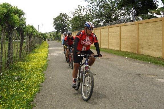 Three cyclists riding on a narrow road lined with greenery and flowers, with a wall in the background. The first cyclist is wearing a red and black jersey and a helmet, while the other two are slightly behind. The scene is set in a sunny, outdoor environment. Cordillera Central mountain bike trail.