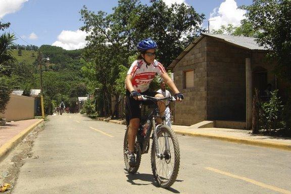 A cyclist wearing a helmet and cycling gear rides a mountain bike down a rural road lined with greenery and small houses. The scene is set on a sunny day, showcasing a picturesque landscape with hills in the background. Cordillera Central mountain bike trail.